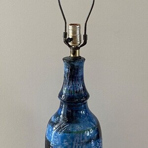 Op de afbeelding: Een blauwe en zwarte keramische tafellamp met een zwarte voet en een zwarte knop. De lamp heeft een wit en blauw abstract ontwerp.