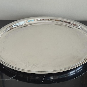 May include: A silver-colored, oval serving tray with a raised, decorative rim. The tray has a reflective surface and appears to be made of metal. The tray is sitting on a black surface.