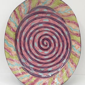 Peut inclure: Une grande assiette ovale en céramique avec un motif peint à la main. Le centre présente un motif en spirale dans des tons de rose et de violet, accentué de tirets turquoise. Le bord est orné de feuilles stylisées dans des tons verts, roses et pêche. L'assiette a une finition brillante.