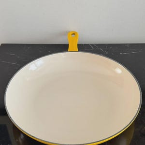 May include: A round, shallow skillet with a bright yellow exterior and a smooth, cream-colored interior enamel coating. The pan features a single, short, yellow handle with a hole at the end for hanging, and a dark gray rim around the edge.