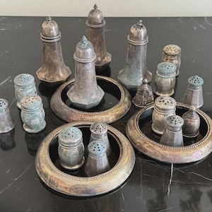 Könnte beinhalten: Eine Auswahl an antiken Salz- und Pfefferstreuern aus Silber. Die Sammlung umfasst verschiedene Formen und Größen, einige mit aufwendigen Details und andere mit schlichtem Design. Die Streuer sind auf einer dunklen Oberfläche angeordnet.