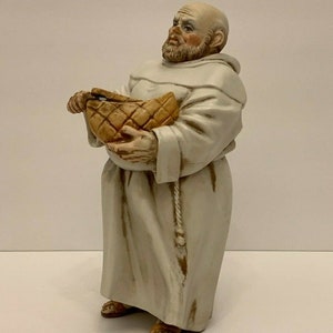Op de afbeelding: Een keramiek beeldje van een monnik die een mand vasthoudt. De monnik draagt een witte robe met een touwgordel. De mand is bruin en heeft een geweven patroon.