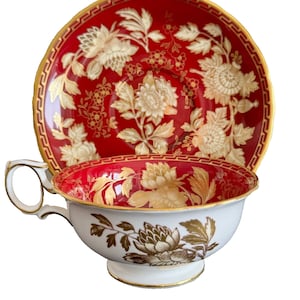 Puede incluir: Un juego de taza y platillo de té rojo y dorado con un patrón floral. La taza y el platillo están decorados con un diseño floral blanco sobre un fondo rojo con detalles dorados.