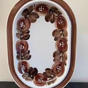 May include: An oval ceramic serving platter with a brown rim and a white center. The platter is decorated with a repeating pattern of brown leaves and red flowers.