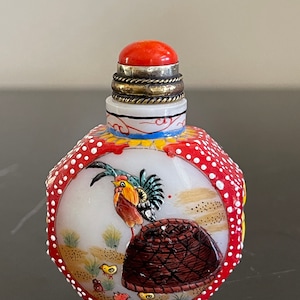 May include: A small, white porcelain snuff bottle with a red coral stopper and a painted scene of a rooster and chicks. The bottle is decorated with red and white dots.