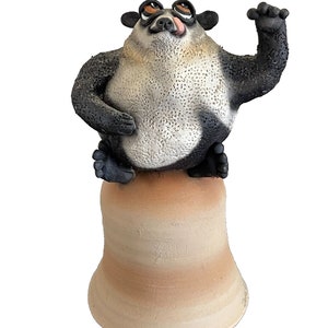 May include: A whimsical figurine of a panda-like creature with a black and white body, perched atop a beige bell-shaped base. The figurine has a playful expression with its tongue sticking out and one arm raised.