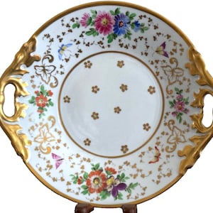 May include: A white porcelain serving dish with gold trim and floral designs. The dish has two handles and is decorated with butterflies and flowers.