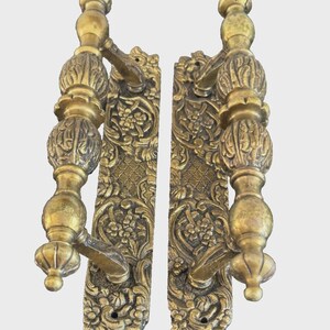 Peut inclure: Deux poignées de porte anciennes en laiton avec des motifs floraux et des volutes ornés. Les poignées ont une surface texturée et détaillée et une teinte dorée chaude. L'ensemble comprend deux poignées, chacune avec une plaque arrière décorative.