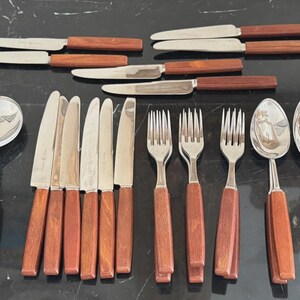 May include: A collection of vintage cutlery with stainless steel blades and tines, and wooden handles. The set includes knives, forks, spoons, and serving spoons. The wooden handles are a dark brown colour.