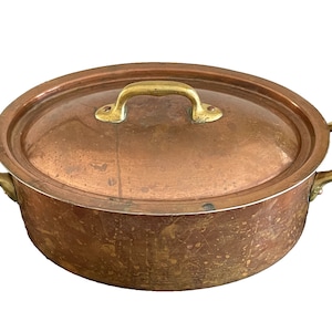 Puede incluir: Una olla de cobre vintage con asas de latón y tapa. La olla tiene una pátina desgastada y envejecida y es de forma ovalada.