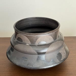 May include: A dark gray ceramic bowl with a wide opening and a rounded base. The bowl features a repeating pattern of semi-circles in a lighter shade of gray. The bowl is sitting on a wooden surface.