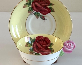 May include: A vintage teacup and saucer set. The cup and saucer are pale yellow with a red rose design. The cup has a pink rose handle and sits on a small wooden stand. Gold trim accents the edges.