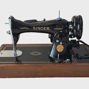 May include: A vintage Singer sewing machine, black with gold detailing, on a wooden base. The machine has a hand wheel, needle, and the Singer logo. The base has a decorative gold border.