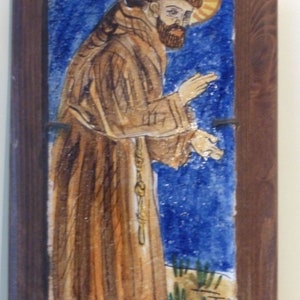 May include: A framed ceramic tile depicting a religious figure in a brown robe with a halo. The figure is standing against a blue background with green plants in the foreground.
