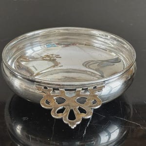 May include: A round, polished silver bowl featuring a single ornate, openwork handle extending from the side. The bowl has a smooth, reflective interior surface and a rounded exterior, showcasing a metallic finish with subtle signs of aging.