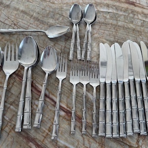 Könnte beinhalten: Eine Sammlung von Edelstahlbesteck mit Bambus-Griffen. Das Set umfasst Löffel, Gabeln und Messer, angeordnet auf einer gemusterten Oberfläche. Das Besteck ist reflektierend und scheint neu zu sein.