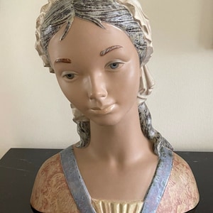 Puede incluir: Un busto de cerámica de una mujer con un gorro blanco y cabello castaño. El busto está pintado en tonos marrones, beige y azul.