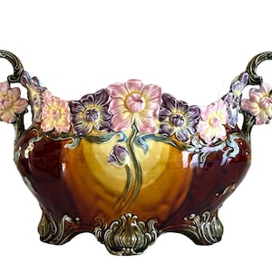 May include: A ceramic decorative bowl with a floral design. The bowl features a mix of colors, including brown, yellow, and pink. It has two handles and is adorned with sculpted flowers. The base has decorative feet.