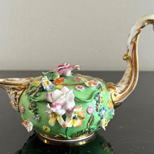 Puede incluir: Lámpara de aceite decorativa de porcelana con base verde y asa curva. La lámpara está adornada con flores y mariposas tridimensionales de colores. Los bordes y el asa están acentuados con detalles dorados, añadiendo elegancia al diseño floral.