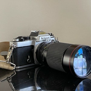 May include: A silver Nikon camera with a black lens attached. The camera has a brown leather strap attached to it.