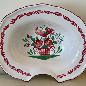 May include: A white ceramic bowl with a red rim and a rooster design. The rooster is painted in red, yellow, and green, and is surrounded by flowers. The bowl has a scalloped edge.