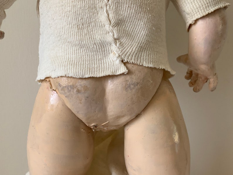 May include: A vintage porcelain doll with a white knitted sweater and a white fabric diaper. The doll has a painted face and is missing some of its paint. The doll is in a standing position with its arms outstretched.