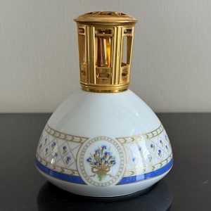 May include: A white ceramic fragrance lamp with a gold-colored metal top. The lamp features a floral design with blue and gold accents. The base has a blue band and a decorative floral medallion.