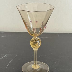 Puede incluir: Una copa de vino de color ámbar con un cuenco hexagonal y un tallo decorativo en espiral. El vaso presenta pequeños acentos rosas en forma de cuentas cerca del borde. La base es redonda y el vaso es de cristal transparente.