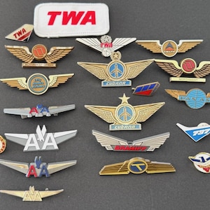 Op de afbeelding: Een verzameling vintage luchtvaartspeldjes en piloteninsignes met diverse logo's waaronder TWA, Delta, Northwest, Pan Am, American Airlines, Cubana, Braniff en Continental. De set bevat metalen gevleugelde badges en een TWA-embleem.