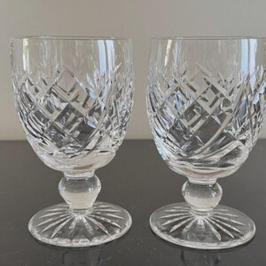 May include: A pair of clear crystal glasses featuring a detailed diamond-cut pattern on the bowl, a rounded stem, and a circular base with a radiating starburst design etched into the glass surface, providing a reflective and textured appearance.