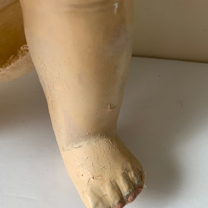 May include: Close-up of a doll's foot made of painted plaster. The foot is painted a pale cream color and has five toes. The toes are slightly curled and the foot is slightly cracked.