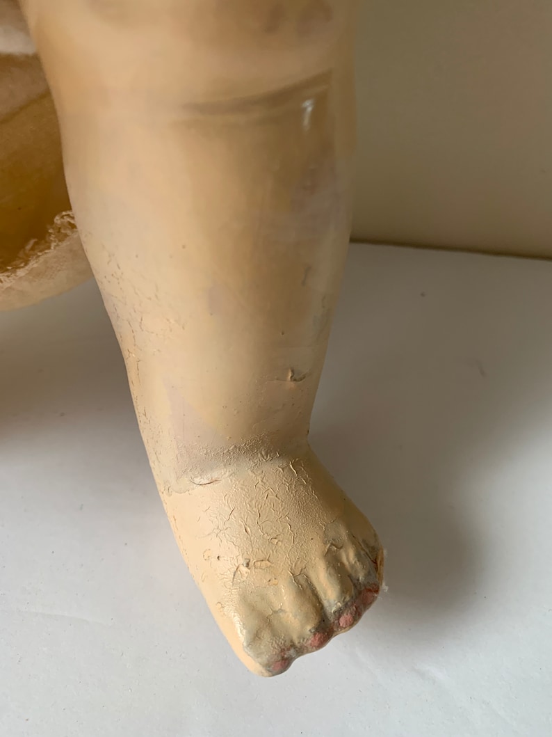 May include: Close-up of a doll's foot made of painted plaster. The foot is painted a pale cream color and has five toes. The toes are slightly curled and the foot is slightly cracked.