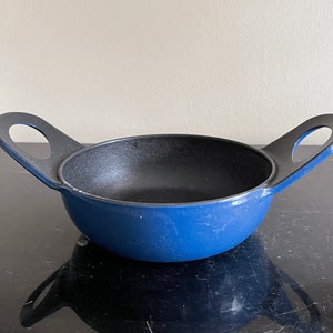 Puede incluir: Un plato de servir redondo de hierro fundido azul con dos asas. El plato es redondo y tiene un interior negro.