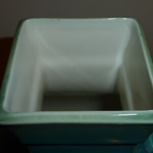 Pode incluir: Um vaso quadrado de cer&acirc;mica com um esmalte verde claro. O vaso tem um interior branco liso e uma borda ligeiramente elevada.