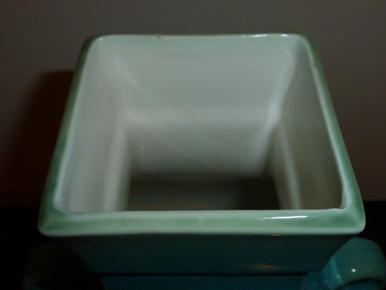 Pode incluir: Um vaso quadrado de cer&acirc;mica com um esmalte verde claro. O vaso tem um interior branco liso e uma borda ligeiramente elevada.