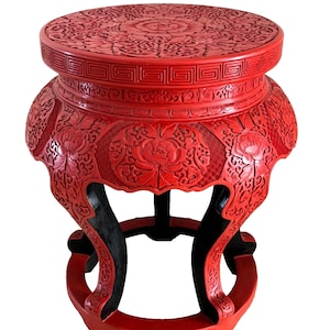 May include: A red, intricately carved wooden stool with a round top and three legs. The stool is decorated with floral patterns and has a black base.