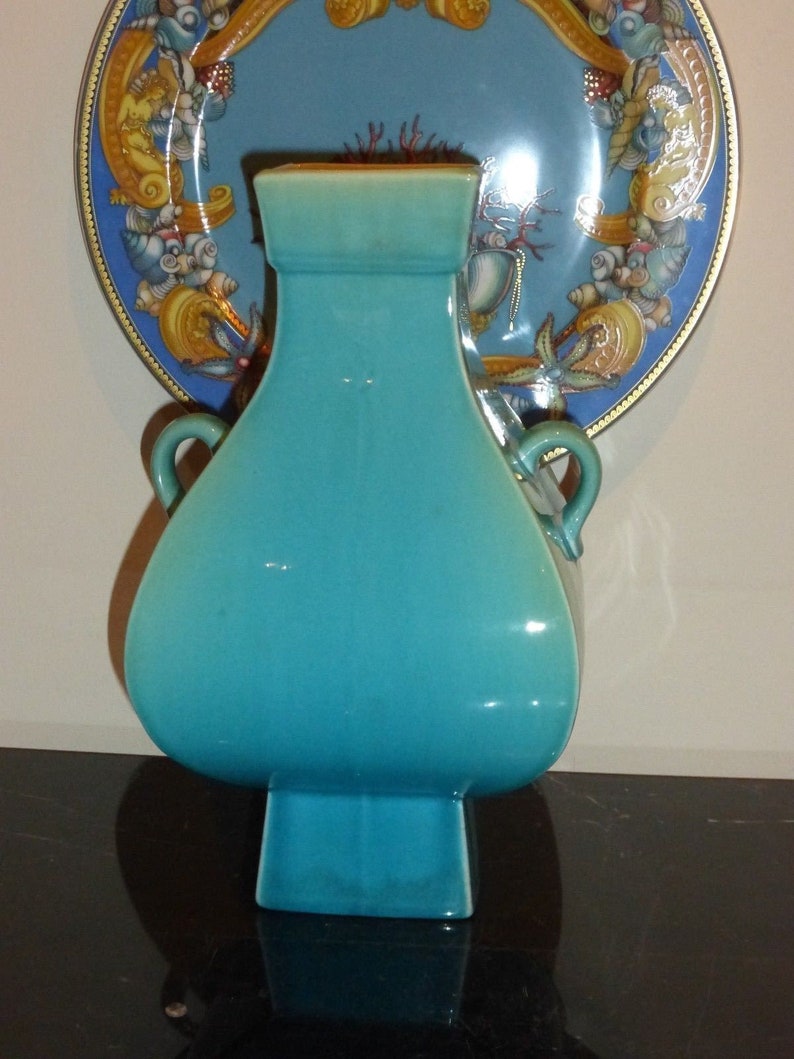 Pode incluir: Um vaso de cer&acirc;mica turquesa com duas al&ccedil;as e uma base quadrada. O vaso &eacute; decorado com um esmalte que lhe confere um acabamento liso e brilhante.