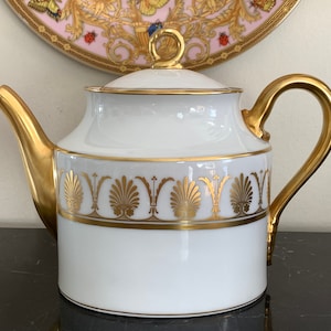 May include: White porcelain teapot with a gold lid and handle. The teapot has a gold band around the middle with a repeating pattern of stylised shells.