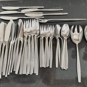 Pode incluir: Uma grande coleção de talheres de aço inoxidável com um padrão linear texturizado nos cabos. O conjunto inclui várias facas, garfos, colheres e dois utensílios de servir grandes, dispostos em grupo sobre uma superfície escura.