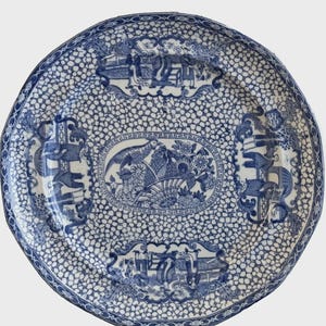 May include: Blue and white ceramic plate with a detailed pattern. The plate features a central oval design with birds and floral elements, surrounded by smaller rectangular scenes. The background is covered in a speckled pattern.