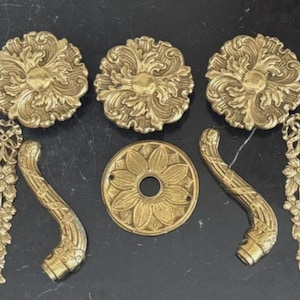 May include: A collection of gold-toned metal hardware components including three large circular medallions with ornate scrollwork, eight elongated floral-patterned vertical strips, two curved handle pieces, and one circular plate with a central hole.