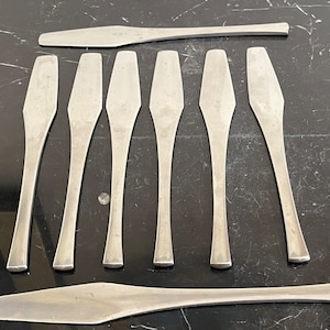 May include: A set of eight silver-toned metal spatulas with rounded edges. The spatulas are arranged in a row on a black surface.