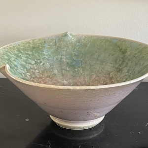 Könnte beinhalten: Eine große, weiße Keramikschüssel mit einem breiten, ausgestellten Rand und einem gewellten Rand. Die Schüssel hat eine hellgrüne und rosafarbene Glasur mit einer strukturierten, gesprenkelten Oberfläche.