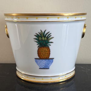 Może przedstawiać: Białe ceramiczne wiadro na lód ze złotą obwódką i uchwytami. Szczegółowa ilustracja ananasa w niebiesko-białej misce ze wzorem znajduje się na środku z przodu. Wiadro ma klasyczny, elegancki design.