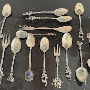 May include: An assortment of antique silver spoons and forks with ornate handles and decorative bowls. The collection includes spoons with twisted stems, detailed figurines, and enamel accents. The pieces are arranged on a dark, reflective surface.