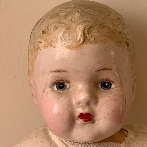 May include: Close-up of a vintage porcelain doll's face. The doll has painted blue eyes, rosy cheeks, and a red painted mouth. The doll's hair is painted a light blonde color.