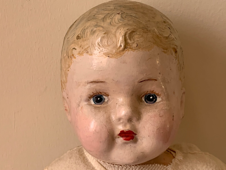 May include: Close-up of a vintage porcelain doll's face. The doll has painted blue eyes, rosy cheeks, and a red painted mouth. The doll's hair is painted a light blonde color.