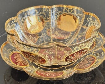 May include: A clear glass bowl and matching saucer set featuring a scalloped edge, gold-painted floral and ribbon motifs, and deep red oval accents. The glass surfaces are decorated with intricate gold filigree patterns and gold-rimmed edges.