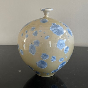 May include: A round ceramic vase with a light beige glaze and blue floral pattern. The vase has a narrow neck and a wide base.