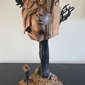 May include: A wooden sculpture of a face with a book and glasses on top of the head. The sculpture is on a wooden base with a black tree trunk.